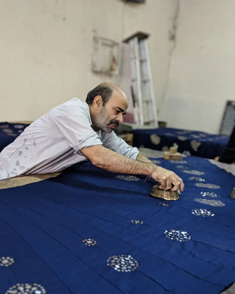 Uma pessoa inclinada sobre uma mesa comprida pressiona um bloco de madeira contra um tecido azul já marcado com motivos circulares brancos